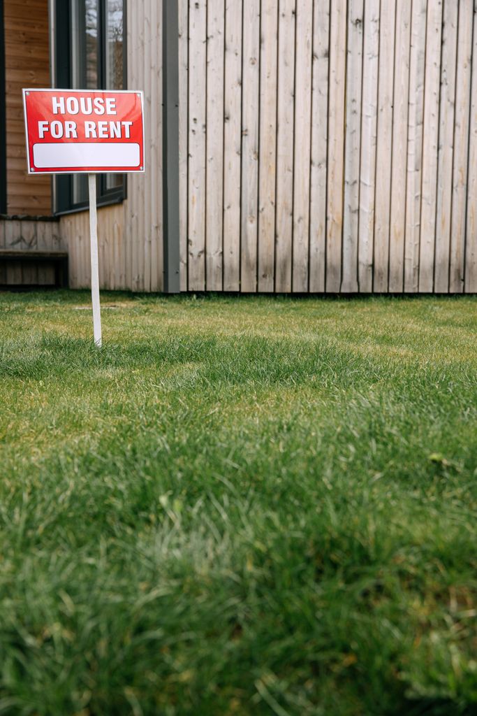 Red 'House for Rent' sign on green lawn with wooden house wall in background.